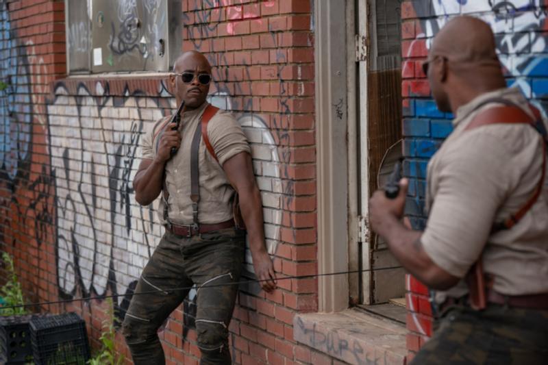 Dwóch mężczyzn w taktycznym stroju i okularach przeciwsłonecznych, trzymających radia, stoi w gotowości w pobliżu pokrytego graffiti ceglanego budynku. Jeden naciska na ścianę, podczas gdy drugi stoi przy otwartych drzwiach, obaj wydają się ostrożni i skupieni.