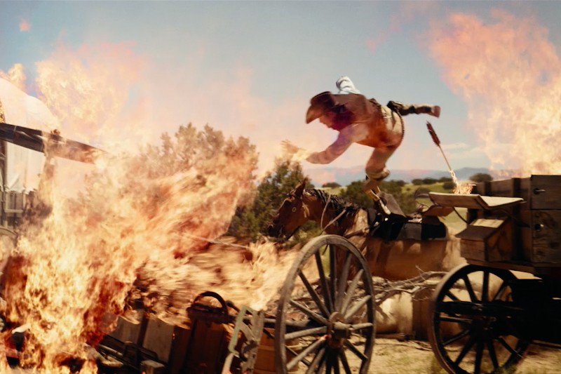 Osoba ubrana w strój kowbojski zostaje wyrzucona w powietrze pośród płonących wozów i intensywnych płomieni, w plenerze w stylu Dzikiego Zachodu pod czystym, błękitnym niebem.