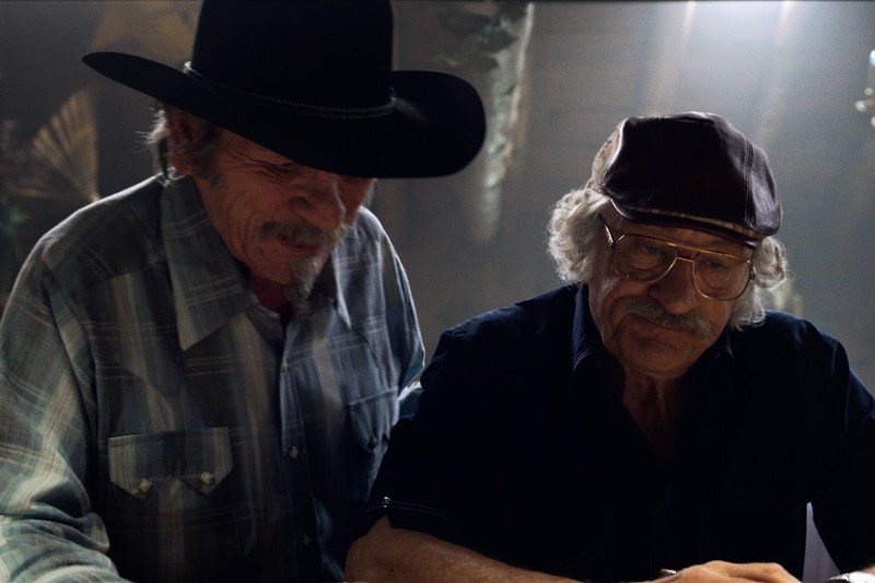 Dwóch starszych mężczyzn z wąsami siedzi obok siebie. Jeden nosi czarny kowbojski kapelusz i kraciastą koszulę, a drugi czapkę, okulary i ciemną koszulę. Wyglądają, jakby byli skupieni na czymś przed sobą.