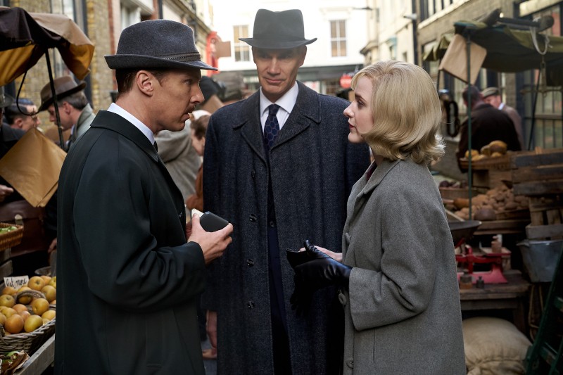 Trzy osoby w ubraniach vintage stoją i rozmawiają na targu na świeżym powietrzu. Dwóch mężczyzn nosi kapelusze i ciemne płaszcze, podczas gdy blondynka w szarym płaszczu i czarnych rękawiczkach słucha. W tle widać stragany na targu i innych kupujących.