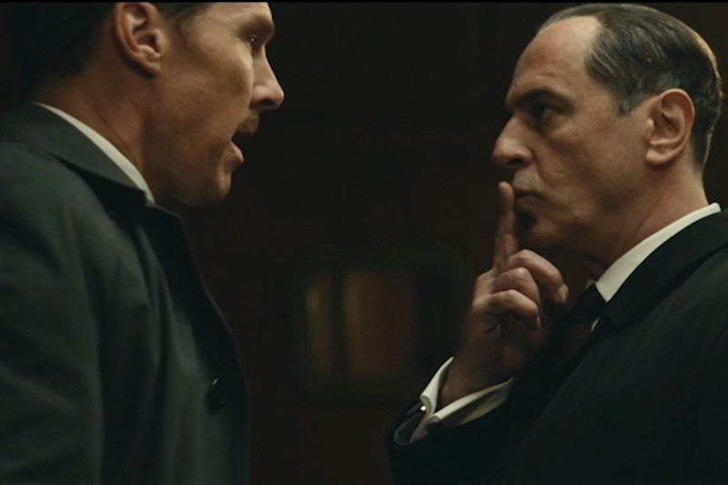 Dwóch mężczyzn w garniturach stoi naprzeciw siebie; jeden z nich przykłada palec do ust, dając znak „cisza”, podczas gdy drugi przygląda mu się uważnie z poważnym wyrazem twarzy.