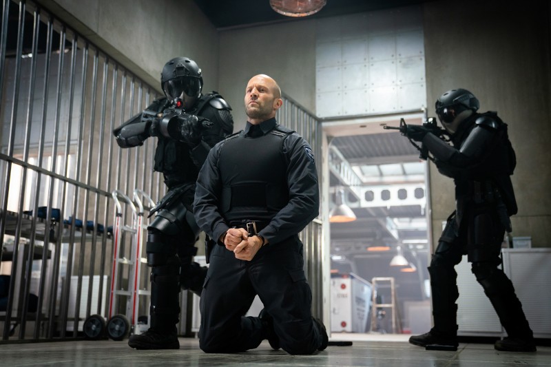 Mężczyzna w czarnym stroju taktycznym klęczy z rękami skute z przodu, podczas gdy dwóch uzbrojonych żołnierzy w czarnych zbrojach mierzą do niego z broni w nowoczesnym, industrialnym pomieszczeniu z metalowymi kratami i sprzętem.