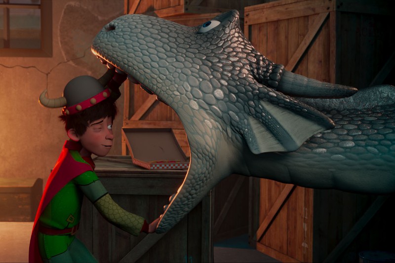 Uśmiechnięty chłopiec w kostiumie stoi z ręką w otwartej paszczy dużego, przyjaznego niebieskiego dinozaura w pokoju wypełnionym drewnianymi skrzynkami, co sugeruje, że między nimi panuje radosna interakcja.