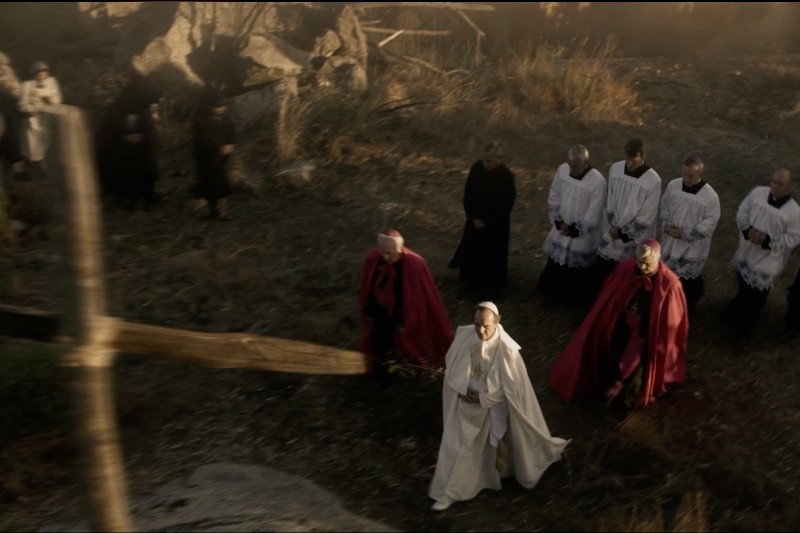 Grupa duchownych w ceremonialnych szatach odbywa procesję na zewnątrz, w pobliżu dużego drewnianego krzyża, idąc przez skalisty, trawiasty teren w delikatnym, przyćmionym świetle słonecznym.