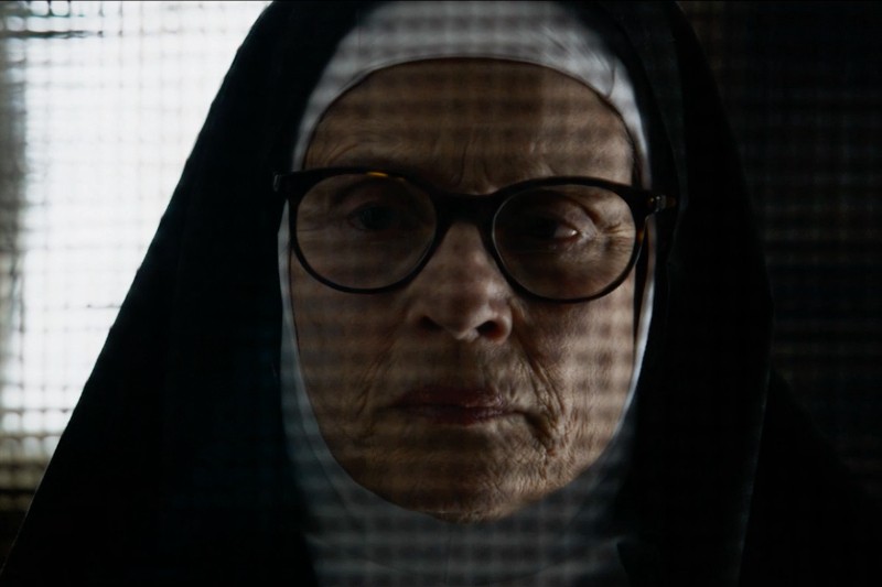 Starsza zakonnica w okularach i czarno-białym habicie jest widoczna przez szklaną kratkę, jej wyraz twarzy jest poważny i zamyślony. Oświetlenie jest słabe, a jej twarz częściowo zacieniona.