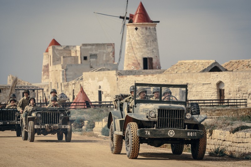 Dwa wojskowe jeepy z żołnierzami w mundurach z II wojny światowej jadą drogą gruntową, mijając zabytkowe kamienne budynki i wiatraki pod zachmurzonym niebem.