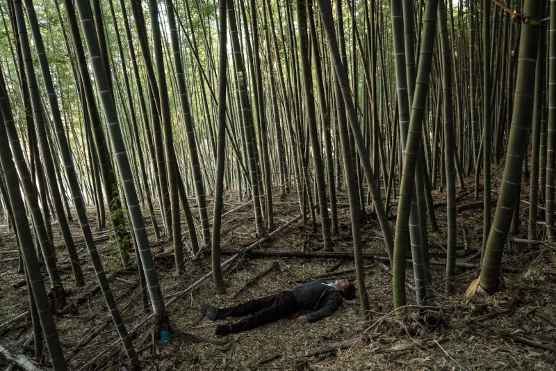 Osoba leży płasko na ziemi w gęstym lesie bambusowym, otoczona wysokimi, prostymi łodygami bambusa i rozrzuconymi suchymi liśćmi. Osoba wydaje się nieruchoma, z zamkniętymi oczami, ubrana w ciemne ubranie.