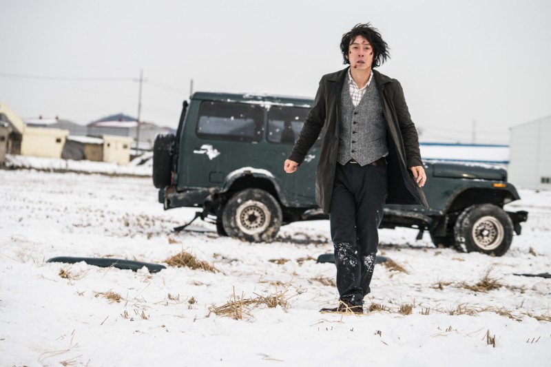 Mężczyzna w ciemnym płaszczu i kamizelce idzie przez śnieg przed samochodem terenowym z uszkodzoną tylną oponą. Scena wydaje się zimna i napięta, w tle widać budynki.