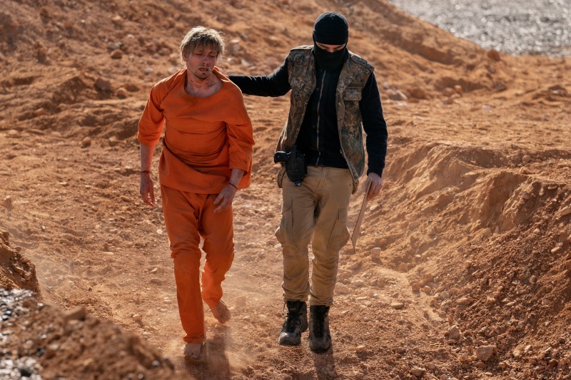 Bosonogi mężczyzna w pomarańczowym stroju, wyglądający na zestresowanego i zaniedbanego, jest prowadzony przez uzbrojonego, zamaskowanego osobnika w wojskowym stroju przez zakurzony, jałowy krajobraz.