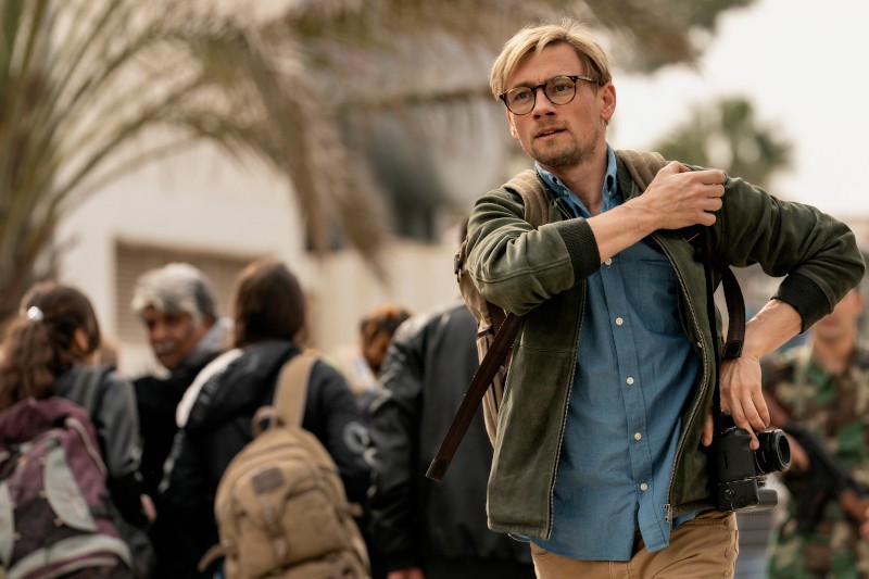 Mężczyzna o blond włosach i okularach, ubrany w zieloną kurtkę i niebieską koszulę, trzyma aparat i patrzy przed siebie, spacerując na zewnątrz w zatłoczonym miejscu. W tle widać kilka osób z plecakami.