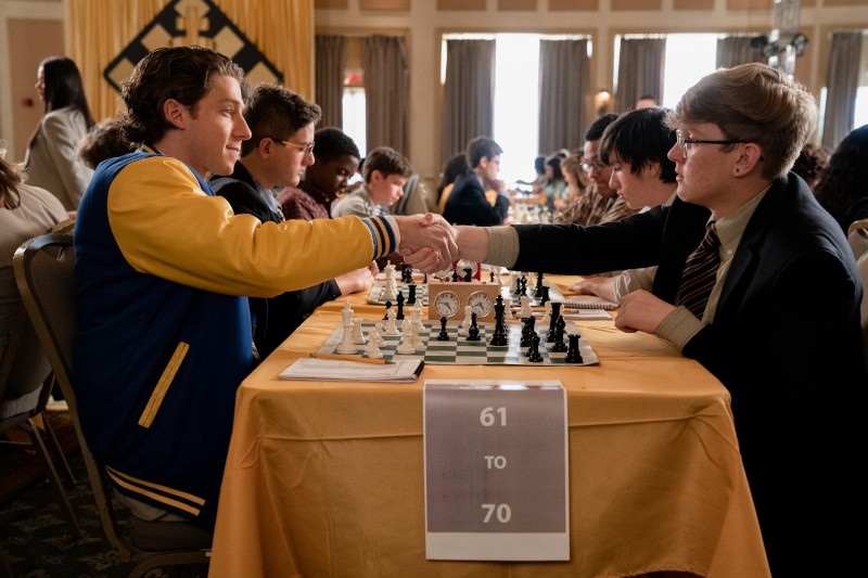 Dwóch młodych mężczyzn ściska sobie dłonie nad szachownicą podczas turnieju. Siedzą przy stole z żółtym obrusem, otoczeni innymi graczami. Na tablicy z przodu widnieje napis 61 do 70. Widzowie i uczestnicy wypełniają salę.