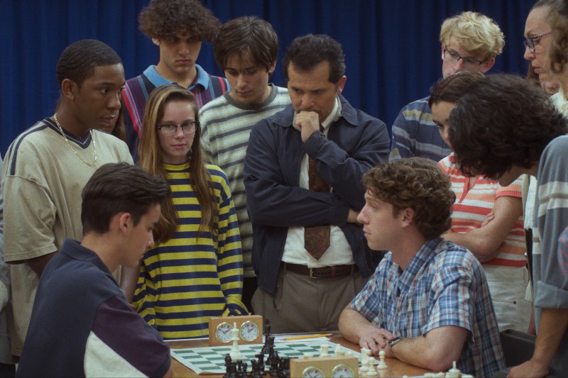 Grupa młodych ludzi i dorosły stoją blisko szachownicy, obserwując dwóch mężczyzn intensywnie grających w grę. Atmosfera wydaje się napięta i skupiona. W tle widać niebieską zasłonę.