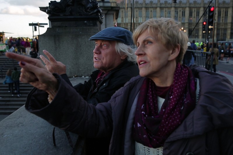 Starszy mężczyzna i kobieta stoją przy kamiennej balustradzie, oboje wskazując i patrząc w dal. W tle widać duży zabytkowy budynek i spacerujących ludzi, co sugeruje miejskie, być może turystyczne otoczenie.