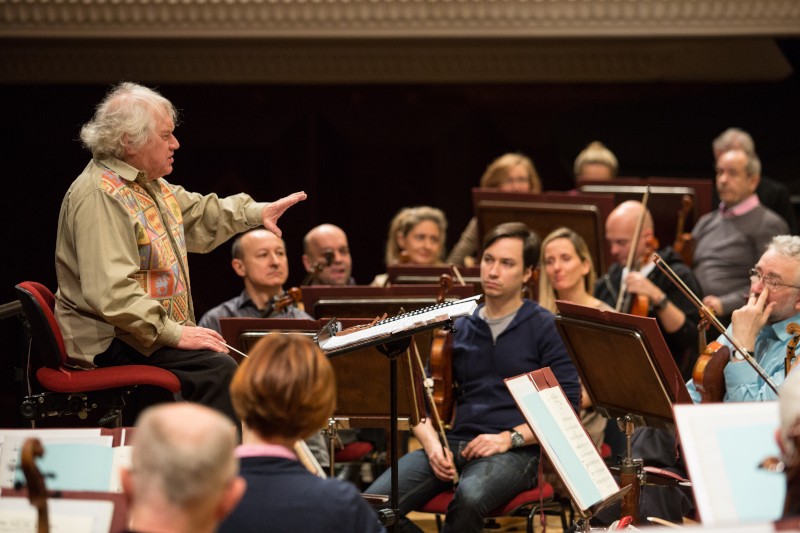 Starszy mężczyzna z siwymi włosami dyryguje orkiestrą, siedząc na krześle i podnosząc rękę, podczas gdy muzycy z instrumentami uważnie obserwują i słuchają na scenie.