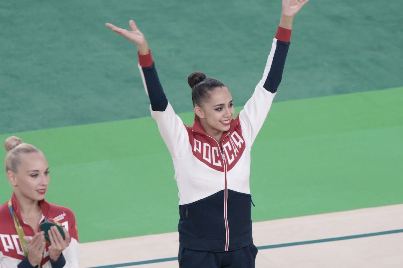 Gimnastyczka w dresie z napisem ROSJA napisanym cyrylicą podnosi ręce i uśmiecha się na podłodze sali gimnastycznej, podczas gdy inna gimnastyczka stoi nieopodal, trzymając medal.