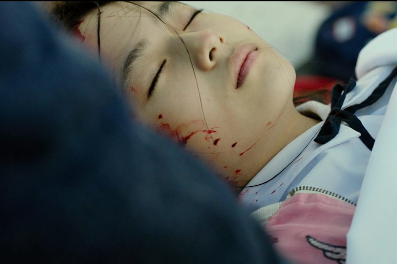 Młoda dziewczyna z zamkniętymi oczami leży na boku, jej twarz i szyja są pokryte krwią. Ma na sobie białą i różową koszulkę, a jej wyraz twarzy jest spokojny, pomimo widocznych obrażeń.