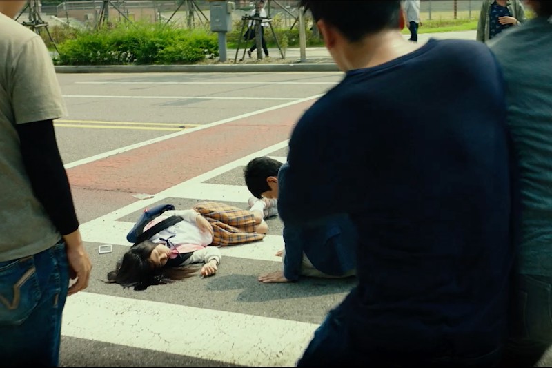 Młoda kobieta leży nieruchomo na przejściu dla pieszych, z kałużą krwi w pobliżu głowy. Mężczyzna klęczy obok niej, a kilka osób stoi w pobliżu, wyglądając na zaniepokojonych. Scena wydaje się napięta i pilna.