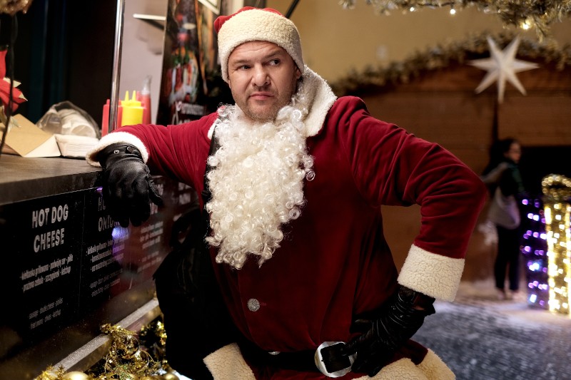 Mężczyzna ubrany w kostium Świętego Mikołaja opiera się o menu food trucka, ma na sobie czarne rękawiczki i patrzy w bok. W tle widać świąteczne światła i świąteczne dekoracje.