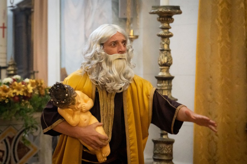 Mężczyzna w długiej, siwej peruce i z brodą, ubrany w żółto-brązową szatę, trzyma figurę dziecka w koronie cierniowej w kościele udekorowanym kwiatami i ozdobnymi świecznikami.