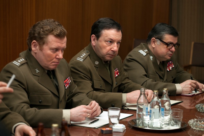 Trzech mężczyzn w mundurach wojskowych z medalami siedzi przy stole konferencyjnym i pisze notatki. Na stole znajdują się butelki z wodą, szklanki i papiery. Otoczenie wydaje się formalne i oficjalne.