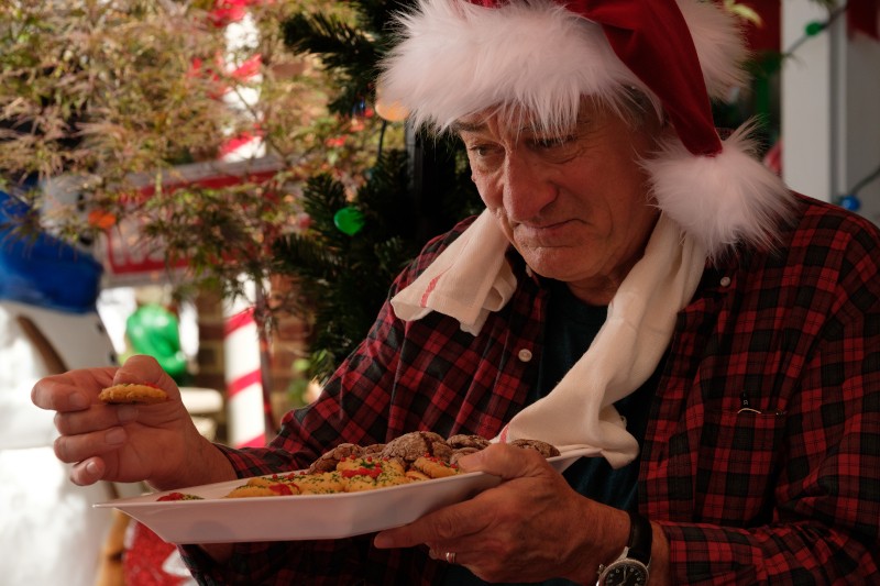 Starszy mężczyzna ubrany w czapkę Mikołaja i kraciastą koszulę trzyma talerz ozdobionych świątecznych ciasteczek i przygląda się im z namysłem, otoczony świątecznymi dekoracjami.