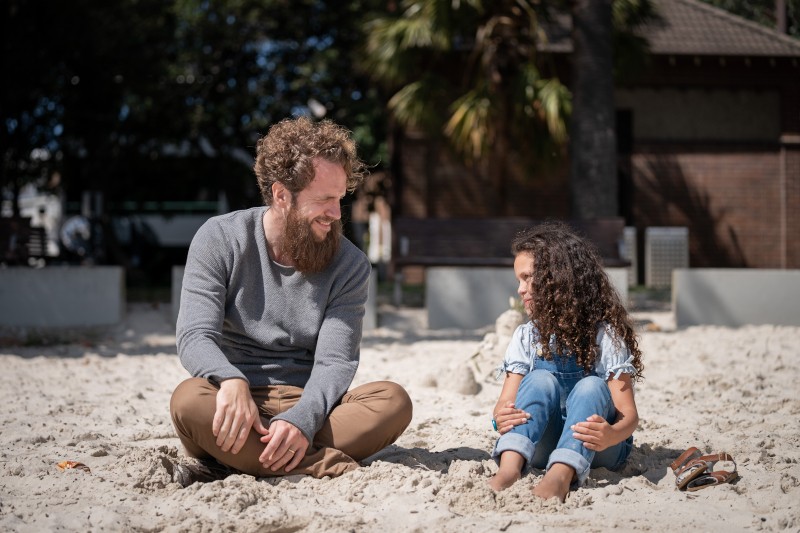 Mężczyzna i mała dziewczynka siedzą boso na piaszczystym podłożu, uśmiechają się i rozmawiają, w tle widać drzewa i budynek.