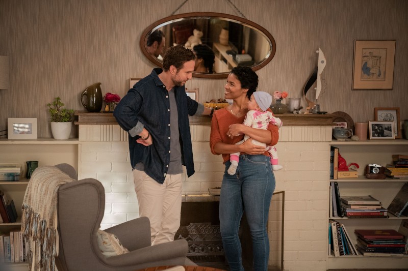 Mężczyzna i kobieta stoją w przytulnym salonie przy kominku; kobieta trzyma niemowlę ubrane na różowo. Oboje dorośli uśmiechają się do siebie ciepło. Pokój jest udekorowany książkami, roślinami i lustrem nad kominkiem.