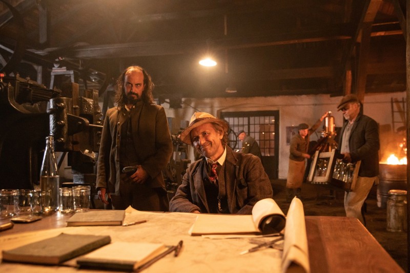 Kilku mężczyzn w staromodnych ubraniach znajduje się w słabo oświetlonym warsztacie. Jeden mężczyzna siedzi i uśmiecha się przy biurku z papierami, podczas gdy inni pracują w tle w pobliżu maszyn i pieca, tworząc zabytkową, industrialną atmosferę.