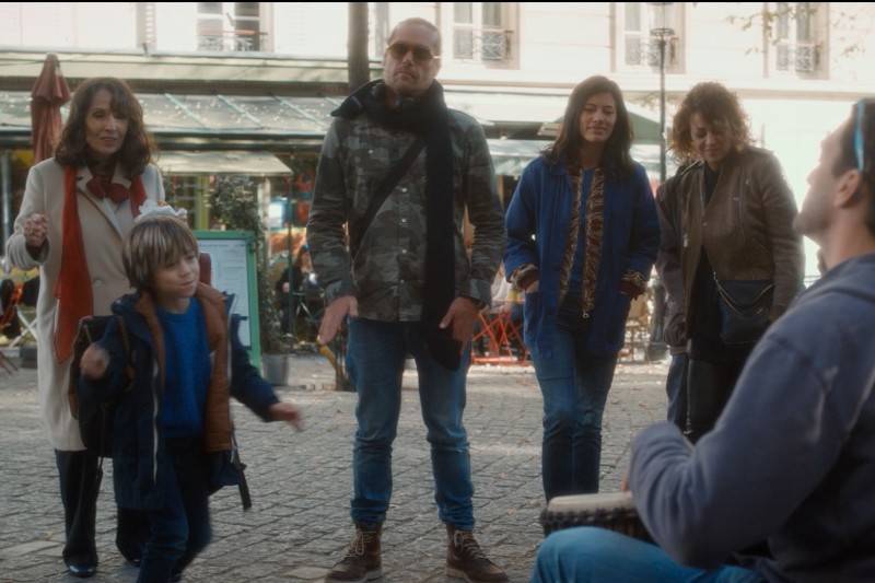 Grupa czterech dorosłych i jedno dziecko stoi na zewnątrz na brukowanej ulicy, słuchając siedzącego muzyka grającego na bębnie. Dziecko wydaje się tańczyć energicznie, podczas gdy dorośli patrzą i się uśmiechają.