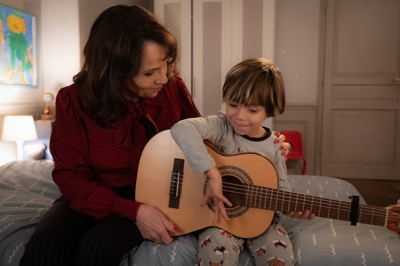 Kobieta siedzi na łóżku obok uśmiechniętego małego dziecka grającego na gitarze akustycznej. Kobieta patrzy na dziecko, które ma na sobie piżamę z psami. Pokój jest delikatnie oświetlony rysunkiem dziecka na ścianie.