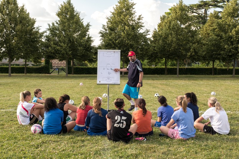 Trener piłki nożnej stoi przy tablicy i wyjaśnia taktykę grupie młodych graczy siedzących na trawie, wszyscy zwróceni twarzą do tablicy, na boisku z drzewami w tle. Piłki nożne są rozrzucone na ziemi.
