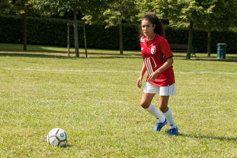 Kobieta w czerwonej koszulce piłkarskiej i białych spodenkach gra na trawiastym boisku, przygotowując się do kopnięcia piłki nożnej. W tle widać drzewa i ławkę.