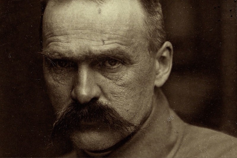 Portret mężczyzny w sepii z bliska, z gęstym wąsem, zmarszczonymi brwiami i intensywnym spojrzeniem, ubranego w koszulę z kołnierzykiem. Obraz ma charakter vintage, historyczny.