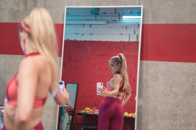 Kobieta w różowym stroju sportowym robi sobie selfie w lustrze na siłowni z czerwoną i szarą ścianą. Jej odbicie pokazuje, jak pozuje z telefonem, a w tle widać sprzęt do ćwiczeń.