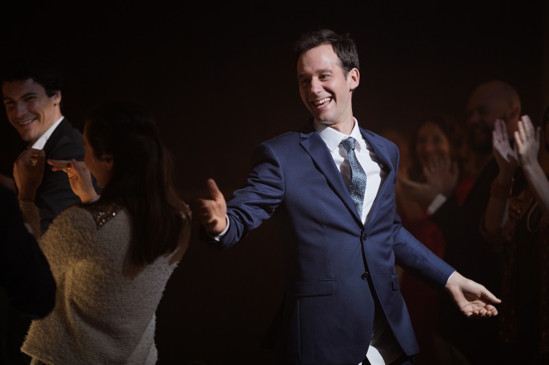 Uśmiechnięty mężczyzna w niebieskim garniturze wyciąga rękę, żeby przybić piątkę, podczas gdy ludzie wokół niego klaszczą i wiwatują w słabo oświetlonym otoczeniu.
