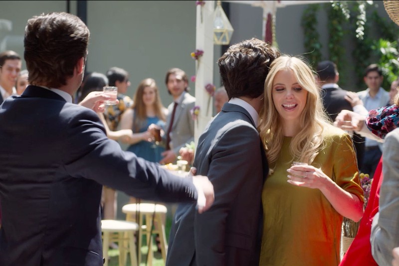 Kobieta w złotej sukni uśmiecha się i trzyma drinka na imprezie na świeżym powietrzu, podczas gdy mężczyzna w garniturze ją przytula. Inni goście są zgromadzeni w tle, wznoszą kieliszki i świętują.