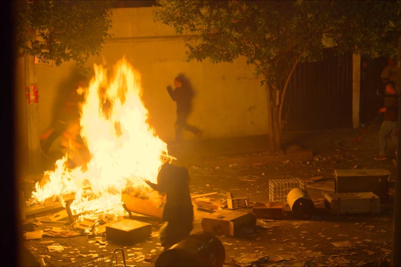 Duży pożar płonie wśród gruzów na miejskiej ulicy nocą. Sylwetki postaci biegną obok, a śmieci i porozrzucane przedmioty pokrywają ziemię. Scena wydaje się chaotyczna i intensywna.