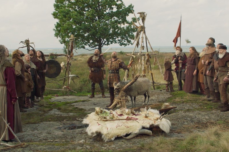 Grupa ludzi w średniowiecznym lub wikingowskim stylu zbiera się na zewnątrz wokół skór zwierzęcych i kości na ziemi, w pobliżu koza i proste drewniane konstrukcje w tle. Widoczne jest duże drzewo i otwarty krajobraz.