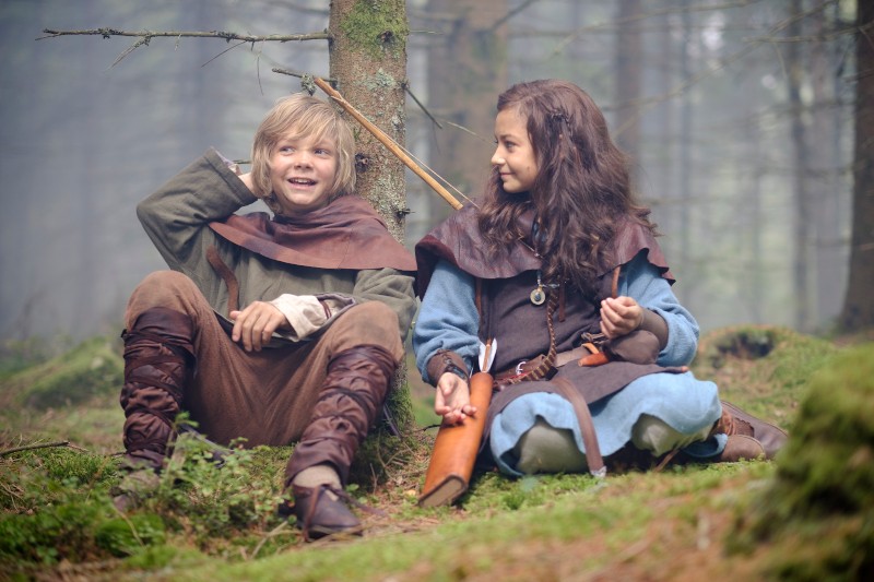 Dwoje dzieci w średniowiecznych strojach siedzi na mchu w lesie. Jedno uśmiecha się i drapie się po głowie, trzymając kokardę; drugie patrzy na niego z małym uśmiechem i trzyma skórzany woreczek.