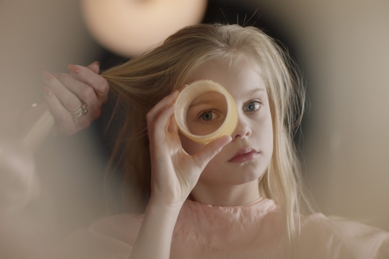 Młoda dziewczyna o długich, blond włosach trzyma wałek do włosów przed okiem i patrzy przez nie, podczas gdy ktoś czesze jej włosy w łagodnie oświetlonym otoczeniu.