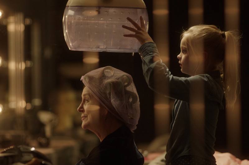 Starsza kobieta z wałkami do włosów siedzi pod suszarką salonową, uśmiechając się, podczas gdy młoda dziewczyna stoi obok niej, dotykając suszarki. Ciepłe oświetlenie tworzy przytulną, intymną atmosferę.