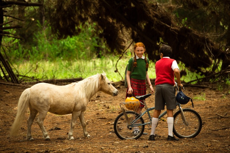 Dwoje dzieci stoi na leśnej polanie z małym białym kucykiem. Jedno dziecko trzyma rower i kask, a drugie, ubrane w zielony sweter i kraciastą spódnicę, uśmiecha się do nich. Światło słoneczne przesącza się przez drzewa.