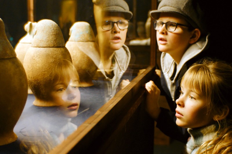 Dwoje dzieci, chłopiec w okularach i kapeluszu oraz dziewczynka o blond włosach, uważnie przyglądają się starożytnym ceramicznym dzbanom wystawionym w szklanej gablocie, w której odbijają się ich twarze.