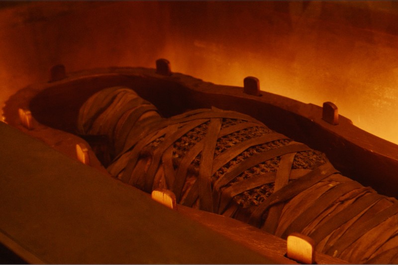 Mumia owinięta brązowymi bandażami leży wewnątrz drewnianego, otwartego sarkofagu, otoczona ciepłym, pomarańczowym oświetleniem.