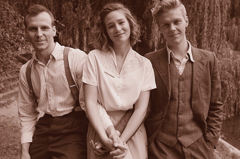Trzech młodych dorosłych z lat 40., dwóch mężczyzn i jedna kobieta, stoją blisko siebie na zewnątrz, uśmiechając się do aparatu. Zdjęcie ma sepiowy odcień, przywołujący nostalgiczną, starodawną atmosferę.