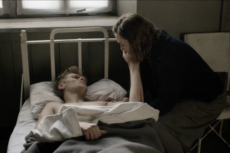 Kobieta siedzi obok mężczyzny leżącego na szpitalnym łóżku, delikatnie dotykając jego twarzy, gdy on na nią patrzy. Światło z okna delikatnie oświetla ponurą scenę, sugerując troskę i zainteresowanie.
