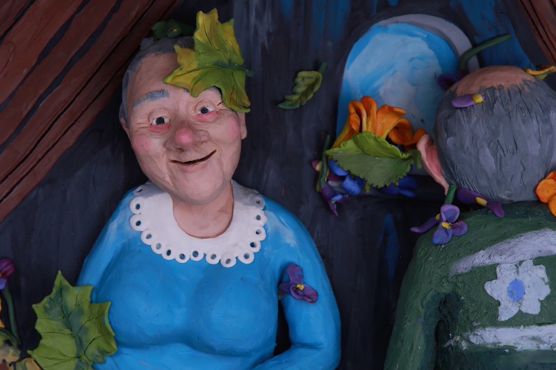 Gliniana rzeźba starszej kobiety w niebieskiej sukience z białym kołnierzykiem, uśmiechnięta. Ma liść na głowie. Obok niej znajduje się inna gliniana figurka z kwiatami i liśćmi, odwrócona tyłem.
