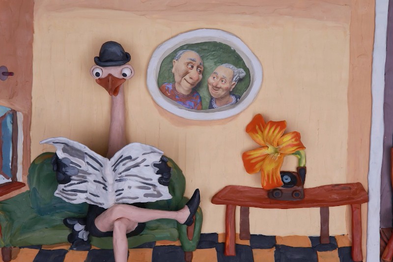 Struś w technice claymation w meloniku siedzi na zielonej kanapie i czyta gazetę. Na stole leży kwiat i aparat, a na ścianie za nim wisi portret dwóch starszych osób.