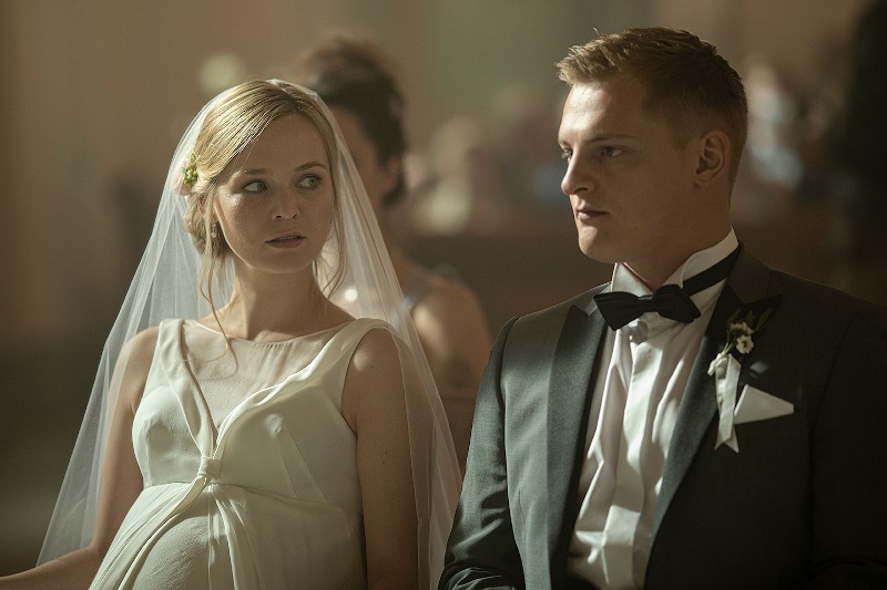 Panna młoda w białej sukni i welonie, widocznie w ciąży, siedzi obok pana młodego w smokingu z butonierką. Patrzą na siebie z poważnymi minami w delikatnie oświetlonym otoczeniu, prawdopodobnie na ceremonii ślubnej.