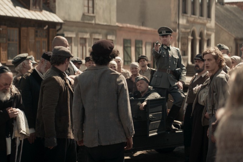 Umundurowany żołnierz w jeepie mierzy z broni do tłumu zdenerwowanych ludzi w staromodnych ubraniach na ulicy o zniszczonych budynkach, co przywołuje napiętą, historyczną scenę.